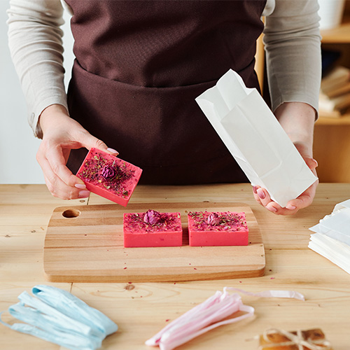 Hands of female holding handmade soap bar and white paper packet over table while preparing gifts for holiday